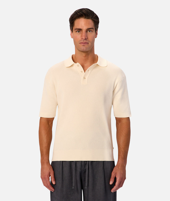 A man wearing a cream-colored short-sleeve knit polo shirt and dark gray drawstring trousers, standing against a light gray studio background.