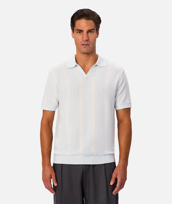 A man wearing a light blue short-sleeve knit polo shirt with subtle vertical stripes, paired with dark gray trousers, against a plain light gray background.