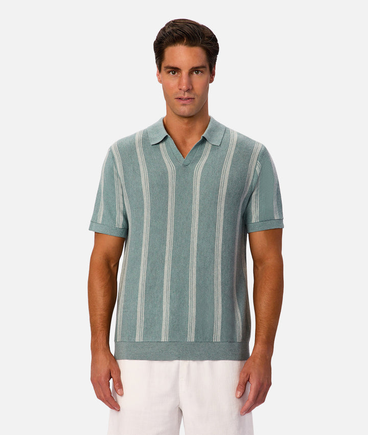 A man modeling a short-sleeve knit polo shirt with vertical stripes in sage green and off-white, paired with white pants, against a light gray studio background.