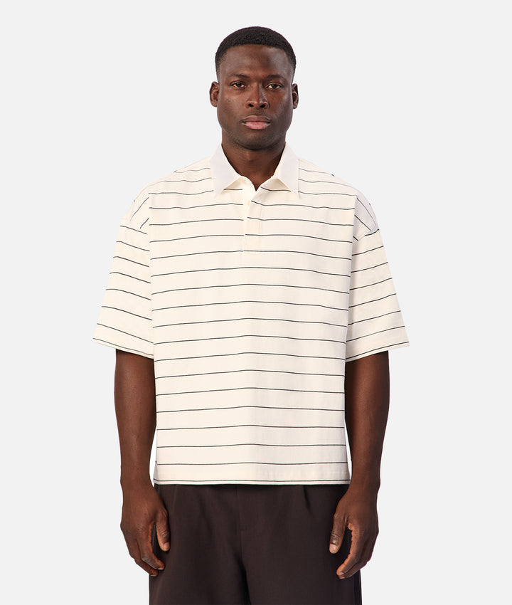 A man wearing a short-sleeve polo shirt with thin horizontal dark stripes on an off-white base, paired with dark trousers, against a light gray background.