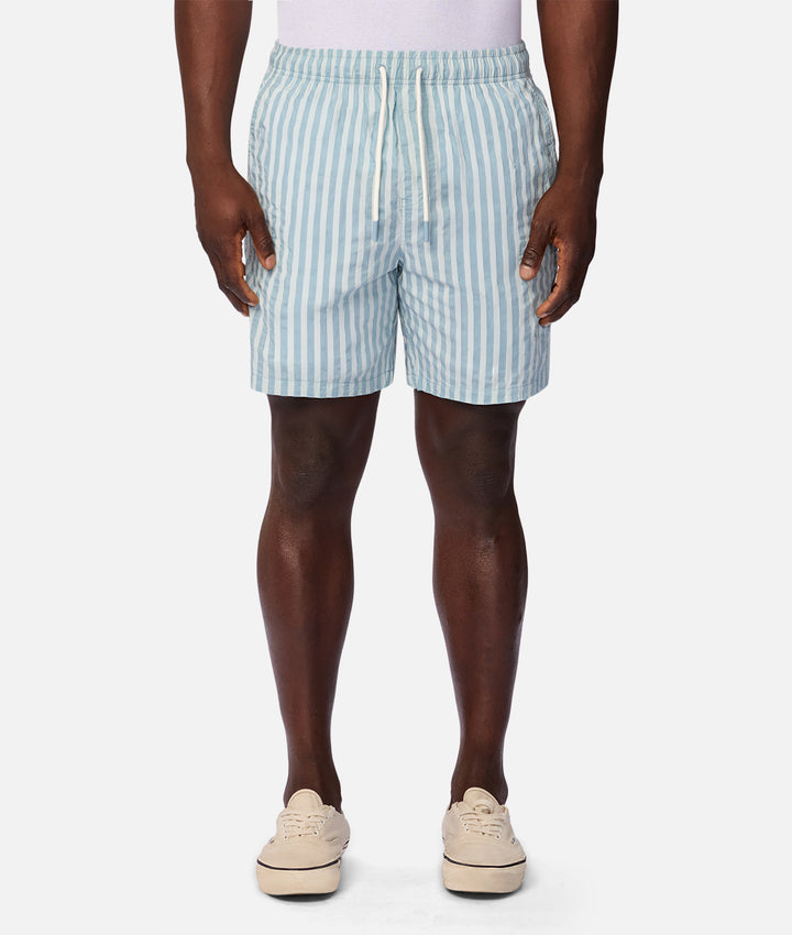 A model from the waist down wearing light blue and white vertically striped swim trunks, paired with beige platform sneakers against a plain white studio background.