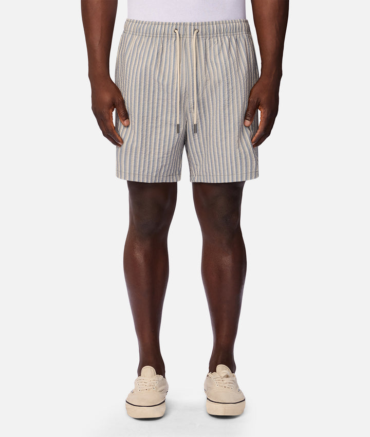 A person wearing light beige and white vertically striped drawstring shorts, paired with cream-colored slip-on shoes, shown from the waist down against a plain light gray studio background.