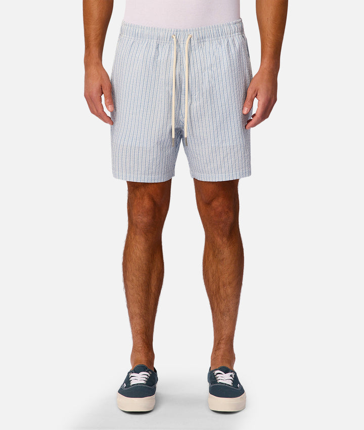 A man wearing light blue and white vertically striped drawstring shorts, paired with a white tank top and navy blue sneakers, standing against a light gray studio background.