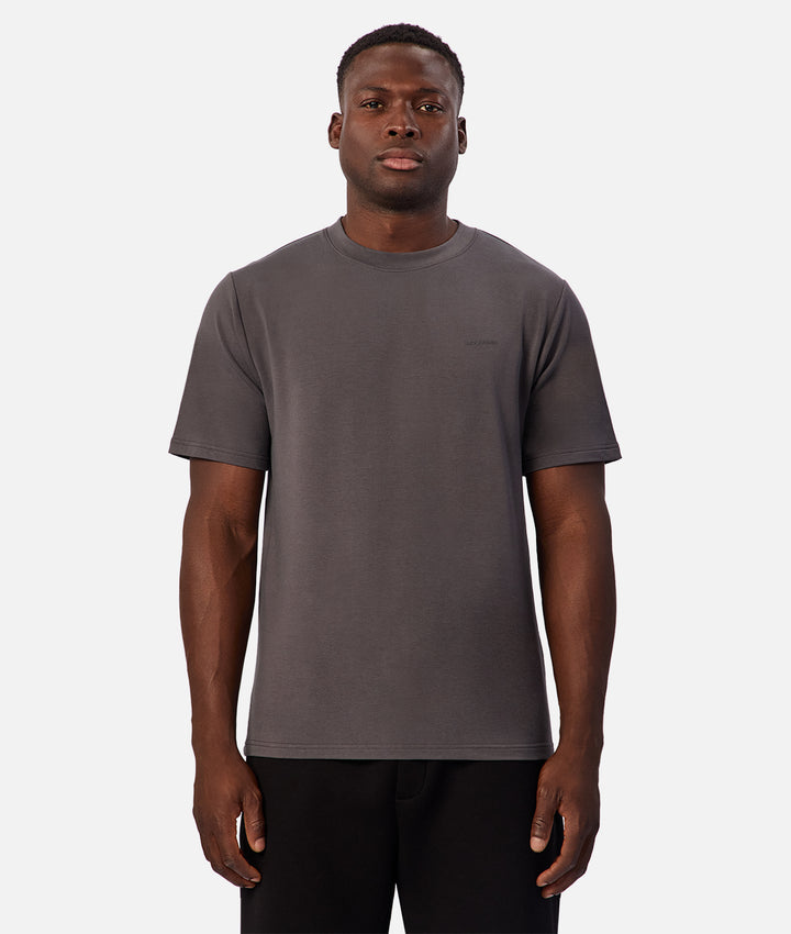 A man wearing a dark gray crew-neck t-shirt and black pants, standing against a light gray background.