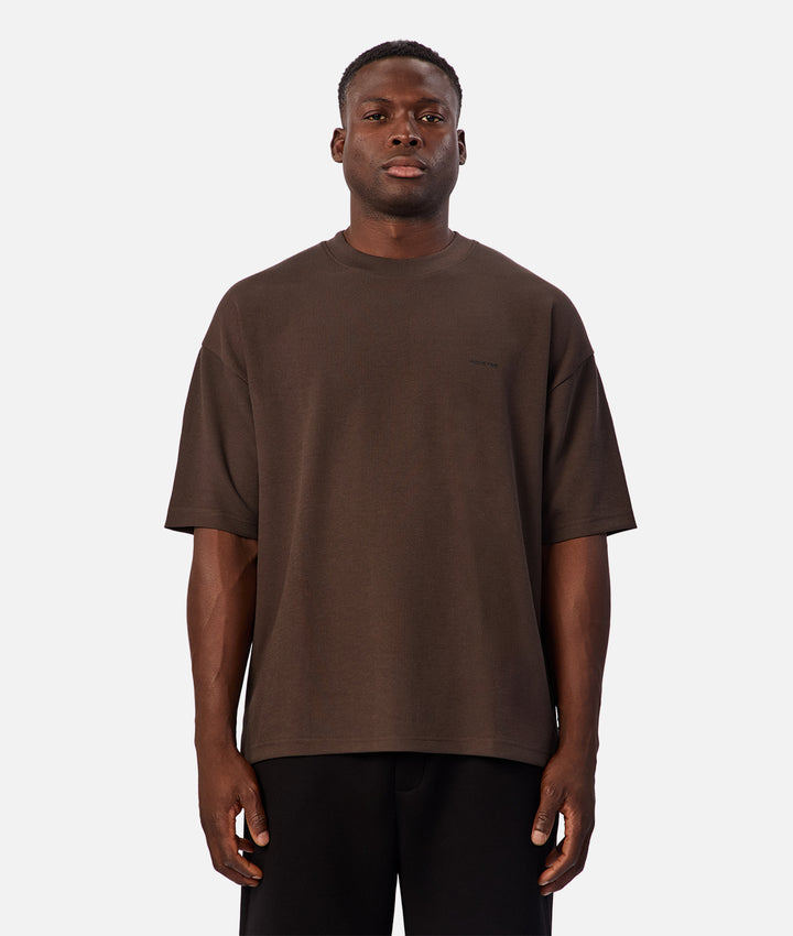 A man wearing an oversized brown short-sleeve crewneck t-shirt and black pants, standing against a light gray background.