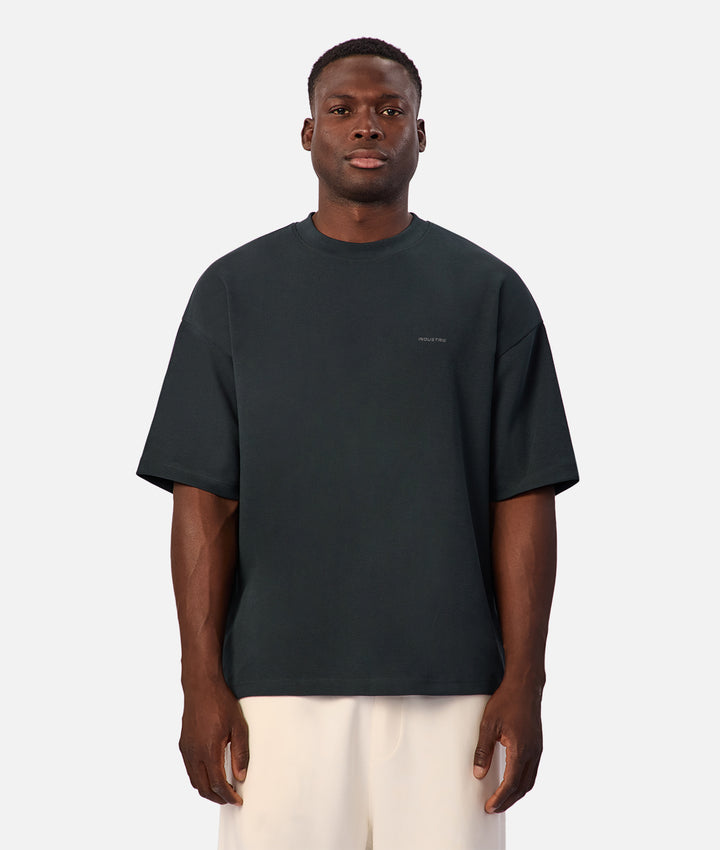 A man wearing an oversized dark charcoal gray t-shirt and off-white trousers, standing against a light gray studio background.