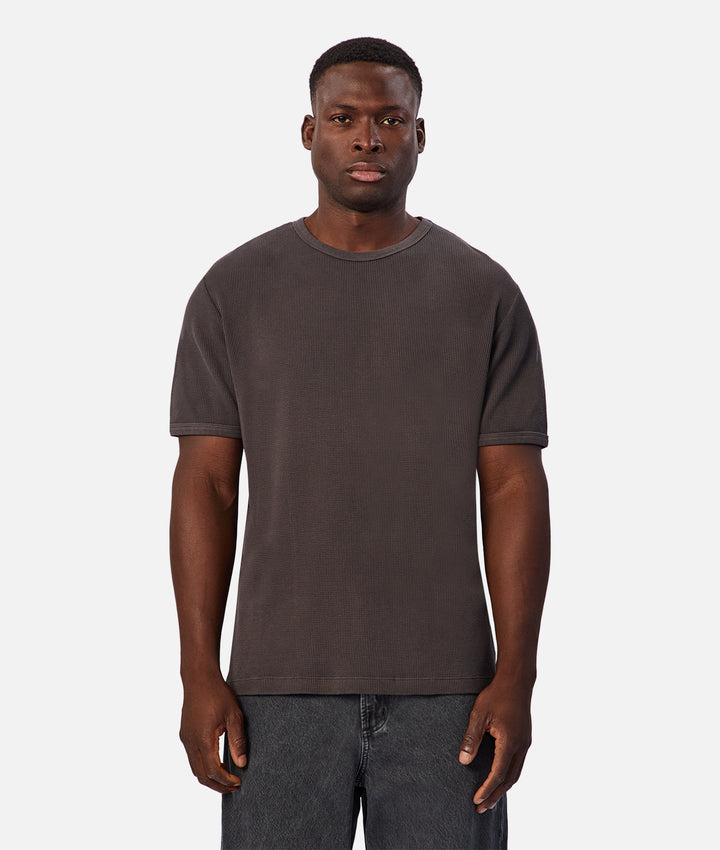 A man wearing a dark brown short-sleeve crewneck t-shirt and dark gray jeans, standing against a light gray background.