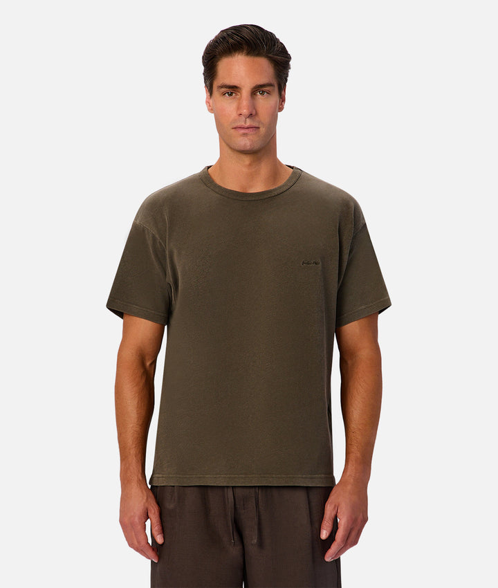 A man wearing a short-sleeve crewneck t-shirt in olive green, paired with dark brown drawstring pants, standing against a light gray studio background.