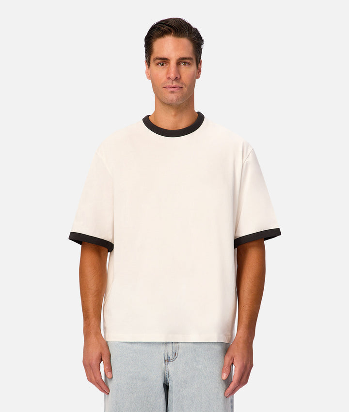 A man wearing a cream-colored ringer t-shirt with black ribbed trim on the crew neck and sleeve cuffs, paired with light-wash jeans, against a light gray studio background.