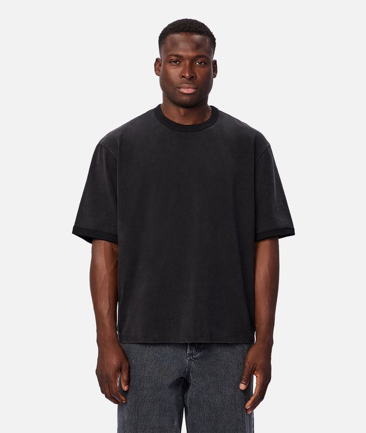 A man wearing an oversized black crewneck t-shirt and gray jeans, standing against a light gray studio background.
