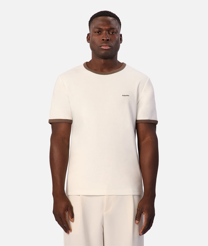 A man wearing a cream-colored short-sleeve t-shirt with brown ribbed trim on the collar and sleeve cuffs, paired with matching cream trousers, against a light gray studio background.