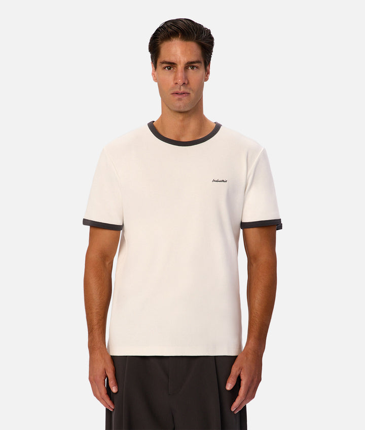 A man modeling a short-sleeve ringer t-shirt in off-white with dark gray trim on the crew neck and sleeve cuffs, featuring a small script logo on the left chest.