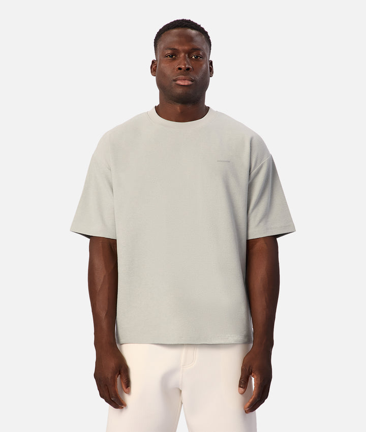 A man wearing a light sage green crewneck t-shirt and off-white trousers, standing against a solid light gray background.