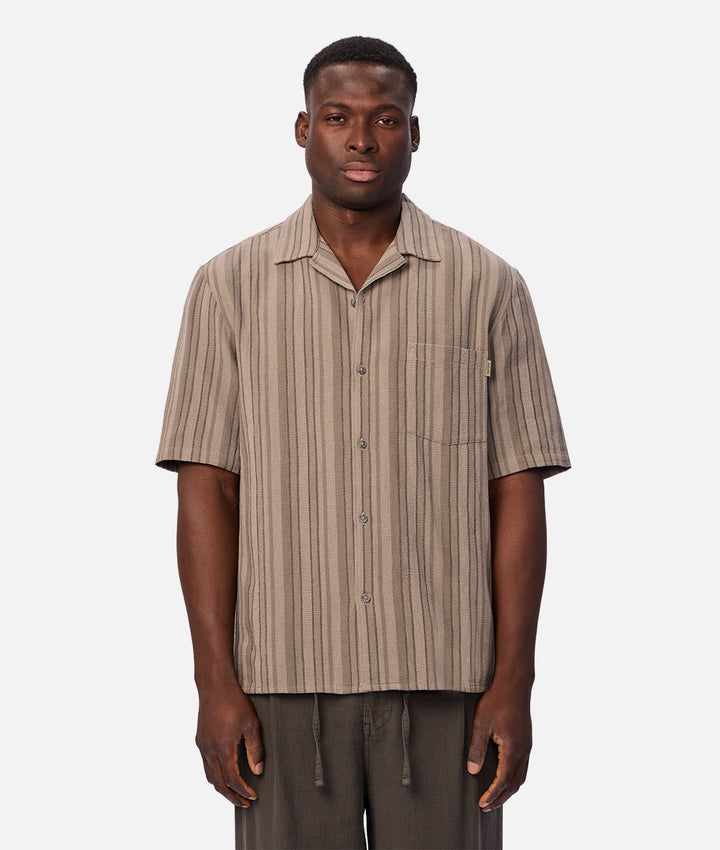 A man wearing a short-sleeve button-up shirt with vertical beige and brown stripes, paired with dark gray drawstring pants, standing against a light gray background.