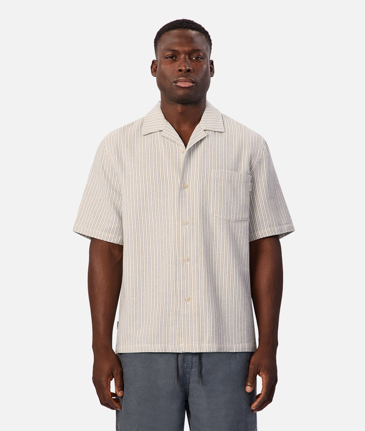 A man wearing a short-sleeve button-up camp shirt with thin vertical stripes in beige and white, paired with gray pants, against a light gray background.
