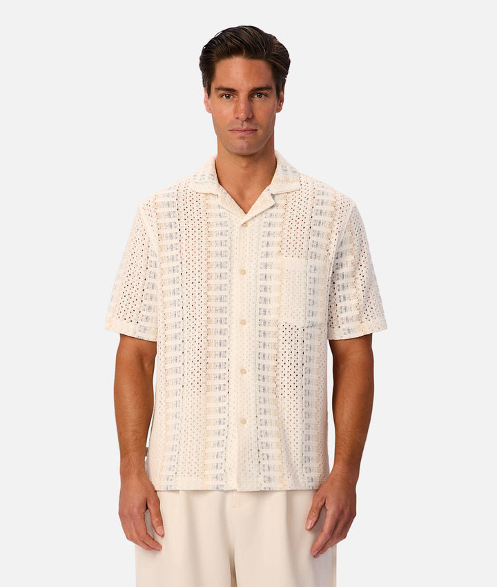 A man wearing a short-sleeve button-up shirt with a textured, open-knit pattern in cream and beige, paired with light beige trousers, against a solid light gray background.