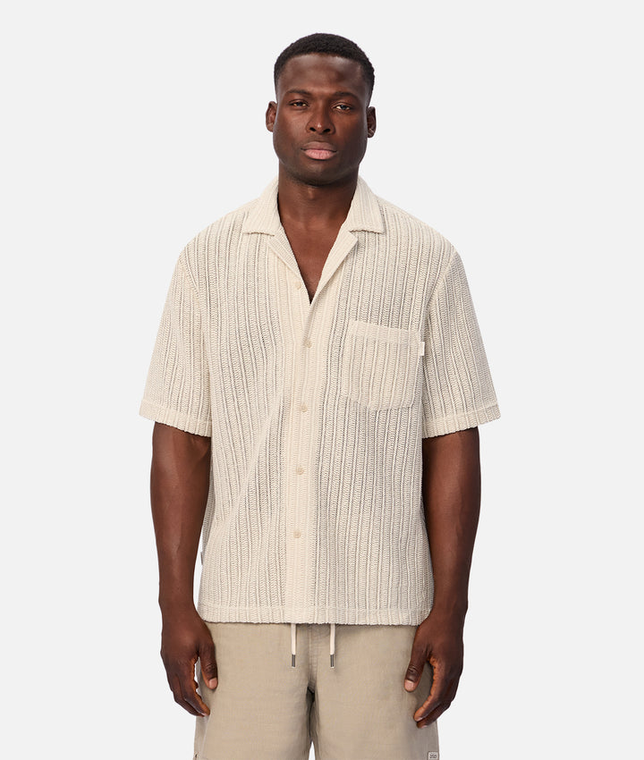 A man wearing a short-sleeve button-up shirt with a textured vertical stripe pattern in cream and beige, paired with beige drawstring pants, against a light gray studio background.
