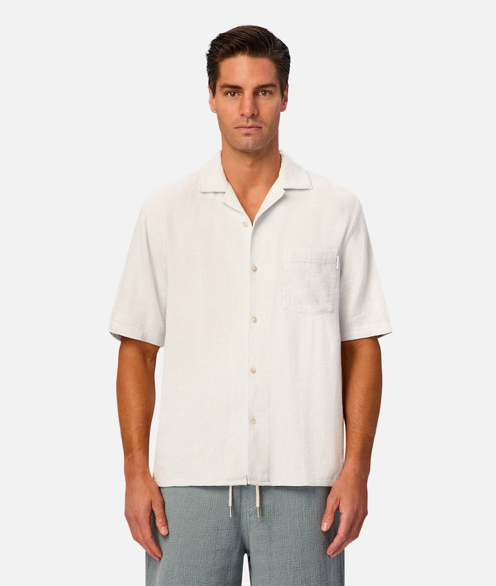 A man wearing a short-sleeve button-up shirt in off-white linen, paired with light gray drawstring pants, standing against a solid light gray background.