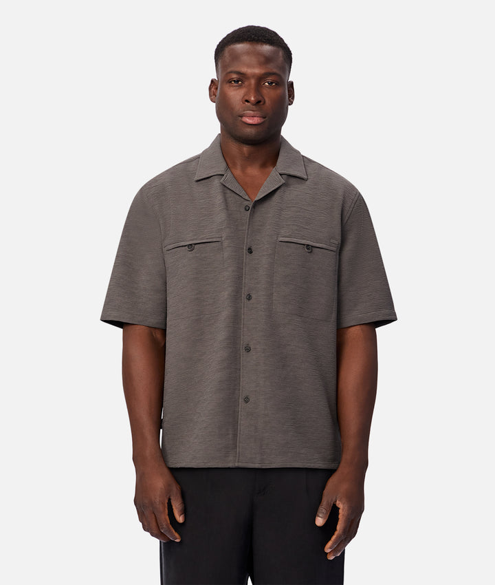 A man wearing a short-sleeve button-up shirt in a heathered dark gray color, paired with black pants, standing against a light gray studio background.