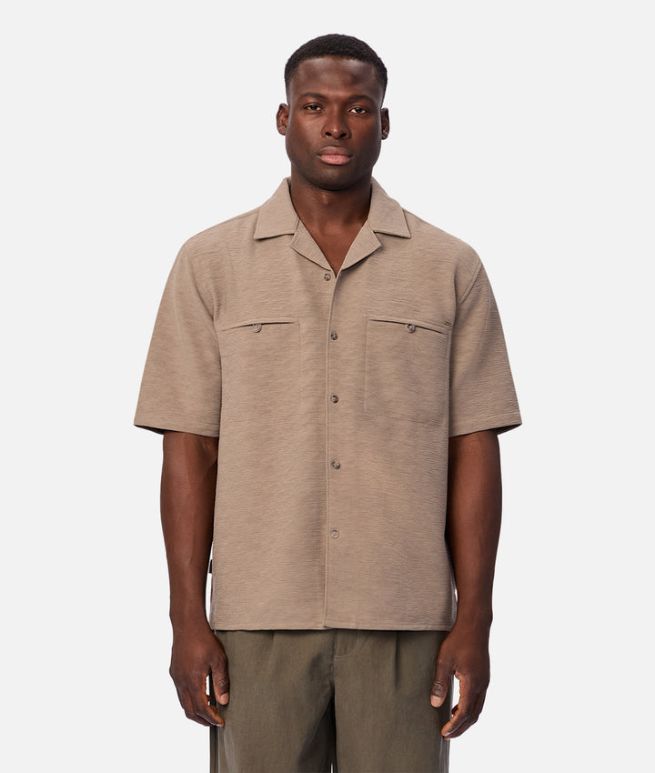 A man wearing a short-sleeve button-up shirt in a light taupe-beige color, paired with olive green trousers, photographed against a solid light gray background.